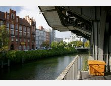 Flussufer mit rotem Backsteingebäude im neugotischen Stil links, flankiert von modernen und klassischen Hausfassaden; im Vordergrund ein eiserner Balkon mit einem orangefarbenen Behälter und einer erhöhten Bahntrasse. Der Fluss ist von grüner Vegetation gesäumt, und im Hintergrund ist eine Brücke zu sehen.