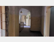 Flur in einem verfallenen Gebäude mit abblätterndem Putz an den Wänden, Holzfußboden und beschädigten Glastüren. Im Hintergrund sind mehrere Türen und eine Deckenlampe zu sehen.