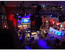 Eine Fernsehstudiokulisse mit einer Bühne im Zentrum, umgeben von roten Vorhängen und Scheinwerfern. Im Vordergrund stehen Tische und Stühle, daneben eine beleuchtete Bar, während technische Ausrüstung und Kameras im Hintergrund zu sehen sind.