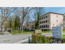 Straßenansicht eines mehrstöckigen Verwaltungsgebäudes mit heller Fassade und dunklen Fensterläden, umgeben von Rasenflächen und Bäumen. Im Vordergrund sind ein Straßenschild mit Adressen, ein Zebrastreifen sowie ein bunt bemalter Stromkasten zu sehen.