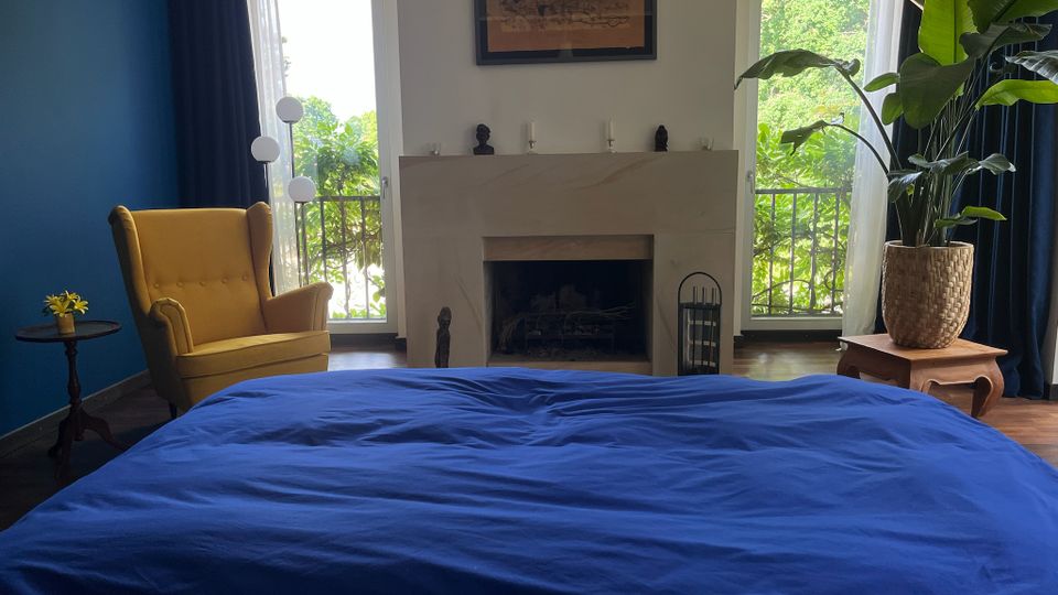 Schlafzimmer mit großem Bett in dunkelblauer Bettwäsche im Vordergrund und einem Kamin mit hellem Steinmantel an der Stirnwand. Links steht ein gelber Ohrensessel vor einer blauen Wand, rechts eine große Zimmerpflanze; zwei hohe Fenstertüren mit Vorhängen öffnen den Blick ins Grüne.