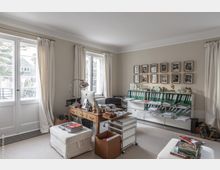 Helles Arbeitszimmer mit naturfarbenen Wänden und großen Fenstern, die reichlich Tageslicht hereinlassen. Der Raum ist eingerichtet mit einem Holztisch, einem Stuhl, mehreren Regalen mit grünen Aktenordnern, Bildern an der Wand und verschiedenen Büro-Accessoires.