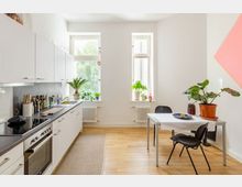 Helle Küche mit weißer Einbauküche, grauer Arbeitsplatte und einem Esstisch mit zwei schwarzen Stühlen. Große Fenster mit Pflanzen auf den Fensterbänken lassen Tageslicht herein, und der Raum hat einen Holzfußboden und eine schlichte Wanddekoration.