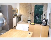 Kleine Küche mit moderner Ausstattung, darunter ein grauer Kühlschrank, ein Backofen, eine Spülmaschine und eine Kaffeemaschine. Im Hintergrund eine unverputzte Ziegelwand und eine grüne Tür mit Graffiti und der Aufschrift „Respect the Neighbours“.