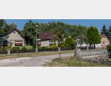 Reihenhäuser mit gepflegten Vorgärten und bunten Blumen, umgeben von Bäumen, entlang einer ruhigen Straße. Im Vordergrund ein Gehweg und ein Zaun aus Metall und Stein.