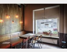 Ein kleines Café mit rustikaler Einrichtung, bestehend aus Holztischen, Holz- und Metallstühlen sowie einer Holzbank. An der Wand hängen freiliegende Glühbirnen, und durch ein großes Fenster mit Fensterbankpflanzen sieht man auf eine Straße mit Gebäuden und einem geparkten Auto.