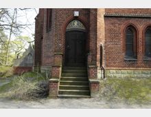 Backsteinfassade mit einem gotischen Eingang einer Kirche oder Kapelle. Der Eingang besteht aus einer braunen Holztür mit verziertem Schmiedeeisen, darüber ein farbiges Mosaik mit christlichem Symbol, flankiert von Spitzbogenfenstern mit Gittern.