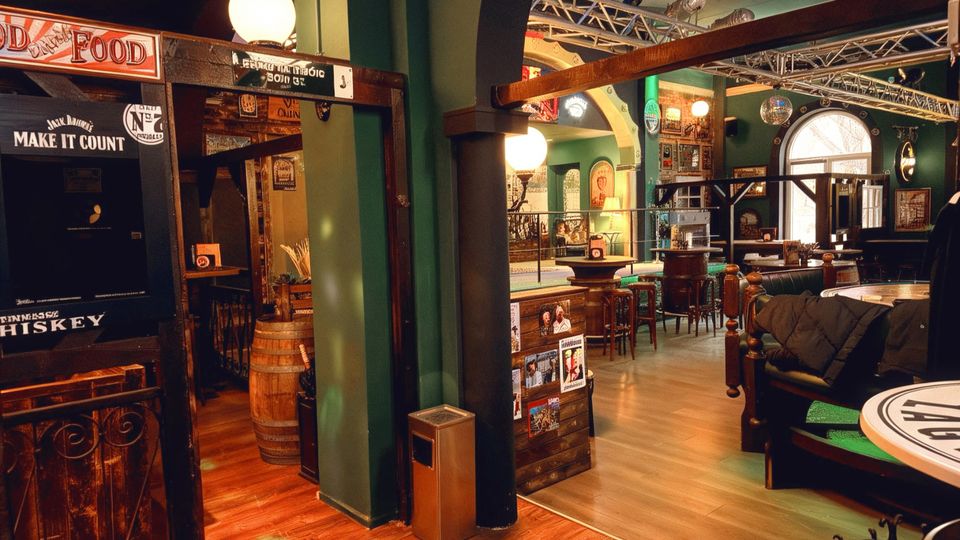 Innenraum einer Bar im Pub-Stil mit grünen Säulen und Rundbögen, Holzfußboden sowie hohen Tischen und Barhockern. An den Wänden hängen zahlreiche Schilder und Poster, und große Lampenkugeln sowie eine Truss-Konstruktion unter der Decke prägen den Raum.