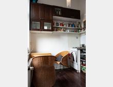 Kleine Küche mit dunklem Holzboden, braunen Hängeschränken, offenen weißen Regalen mit Geschirr und Lebensmitteln, sowie einem Gasherd. Im Vordergrund stehen zwei Rattanstühle und ein kleines Holztischchen an einer weißen Wand.