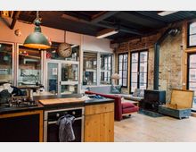 Gemütlicher Loft-Raum mit rustikalem Design, bestehend aus einer offenen Küche mit Holzfronten, einer Sitzgruppe mit Sofas und gemusterten Kissen sowie einer Glaswand mit Blick auf einen Werkstattbereich. Die Backsteinwände, schwarzen Metallfensterrahmen und ein Ofen verstärken den industriellen Charakter.