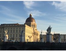 Historisches Gebäude mit einer großen Kuppel im goldenen Licht, umgeben von klassischer Architektur. Im Vordergrund eine Brücke mit mehreren weißen Statuen auf Sockeln sowie historischen Laternen.