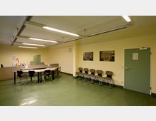 Ein Wartebereich mit gelb gestrichenen Wänden und grünem Boden. Links steht ein Empfangstresen mit Bürostühlen und Computerausrüstung, rechts befinden sich mehrere Stühle an der Wand, darüber hängen zwei Bilder; eine grüne Tür ist ebenfalls sichtbar.