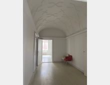 Ein schlichter, weiß gestrichener Raum mit Tonnengewölbedecke und einem Holzboden. Im Hintergrund führt eine geöffnete Tür zu einem weiteren Raum mit einem großen Fenster, durch das natürliches Licht einfällt; rechts stehen zwei Stühle, einer davon rot.