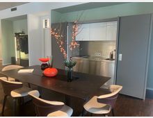 Moderne Küche mit einem Esstisch aus dunklem Holz, umgeben von gepolsterten Stühlen in Creme- und Brauntönen. Im Hintergrund eine Küchenzeile aus Edelstahl mit grauen Schiebetüren; auf dem Tisch befinden sich bunte Vasen und ein Arrangement von Zweigen mit rosa Blüten.