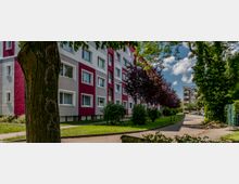 Mehrstöckiges Wohngebäude mit weiß-roter Fassade, flankiert von gepflegten Grünflächen mit Bäumen und Sträuchern. Rechts im Bild verläuft ein Gehweg, der in Richtung weiterer Wohngebäude führt.