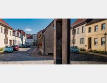Blick auf eine schmale, gepflasterte Altstadtstraße mit niedrigen Wohnhäusern in hellen Fassaden und roten Ziegeldächern. Am rechten Bildrand steht eine spiegelnde Glasfläche, die die gegenüberliegenden Gebäude und geparkte Autos reflektiert.
