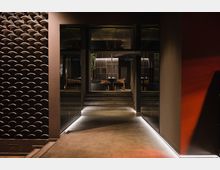 Eingangsbereich eines modernen Restaurants mit beleuchtetem Gang aus Beton, der zu einer erhöhten Ebene mit Tischen und Holzstühlen führt. Links befindet sich eine dunkel strukturierte Wand, rechts eine glatte rote Oberfläche mit indirekter Beleuchtung.