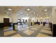 Modernes Eingangsfoyer mit klarer Linienführung, beige-schwarzen Fliesenböden und einer Decke mit rechteckigen, eingelassenen Leuchtelementen. Der Raum umfasst Sicherheitsspinnen, Türen zu angrenzenden Räumen und Informationsschilder.