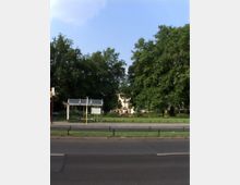 Straßenszene mit einer Bushaltestelle mit gläsernem Unterstand im Vordergrund, dahinter eine Grünfläche mit großen, dicht belaubten Bäumen und vereinzelt sichtbaren Gebäuden im Hintergrund.