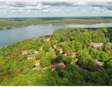 Luftaufnahme einer waldreichen Landschaft mit mehreren Gebäuden, teilweise mit roten Dächern, verteilt zwischen den Bäumen. Im Hintergrund ein großer See, umgeben von dichtem Wald, und ein bewölkter Himmel.