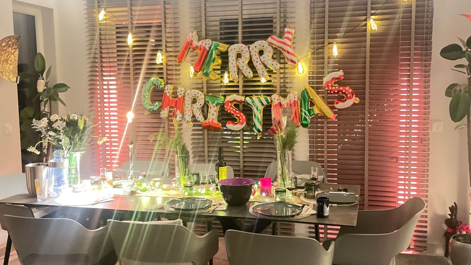 Essbereich in einem modernen Raum mit einem gedeckten Tisch, umgeben von acht grauen Polsterstühlen, dekoriert mit Weihnachtslichtern und einem „Merry Christmas“-Bannerschriftzug vor bodentiefen Jalousien. Pflanzen und dezente Beleuchtung ergänzen das Ambiente.
