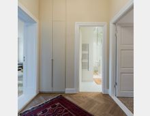 Flur mit hellem Holzboden im Fischgrätmuster, ein roter Teppich liegt auf dem Boden. Zu sehen sind geöffnete Türen: links ein Wohnraum, mittig ein Badezimmer mit Handtuchheizkörper, und rechts ein weiterer geschlossener Raum.