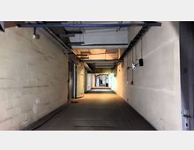 Langer, schmaler Flur in einem Industriegebäude mit blanken, hellen Wänden und sichtbaren Leitungen an der Decke. Auf beiden Seiten befinden sich Türen, und an der rechten Wand hängen verschiedene Schalter und Kabel herunter.