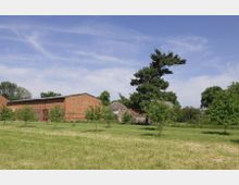 Eine ländliche Szene mit einer großen, roten Backsteinscheune und mehreren kleinen Bäumen auf einer Grasfläche. Im Hintergrund sind weitere Gebäude und ein hoher Baum zu sehen, umgeben von einer klaren, blauen Himmelslandschaft.