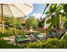 Gartenbereich eines Cafés oder Restaurants mit Holztischen und grünen Stühlen, umgeben von gepflegten Hecken und bunten Blumen. Große Sonnenschirme spenden Schatten, und im Hintergrund sind Bäume sowie ein Gebäude mit roten Ziegeldächern zu sehen.