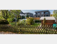 Ein kleiner Garten mit einem Holzzaun im Vordergrund, einer Holzhütte und einem Gewächshaus in der Mitte. Im Hintergrund stehen moderne, dunkelgraue Mehrfamilienhäuser sowie ältere Gebäude, umgeben von Bäumen und Sträuchern.