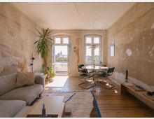 Wohnzimmer in einem minimalistischen Stil mit rohen, verputzten Wänden und Holzdielenboden. Im Vordergrund steht eine helle Couch, daneben Pflanzen und ein runder Esstisch mit vier Stühlen vor einem hellen Fenster mit Blick nach draußen.