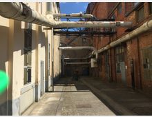 Industrieller Hinterhof mit Backstein- und verputzten Gebäudewänden, verbunden durch mehrere freiliegende Rohre an Metallgestellen. Eine schmale asphaltierte Gasse führt durch den Bereich, umgeben von Fenstern und Türen.