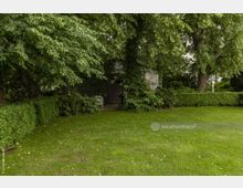 Ein grüner Garten mit kurzgemähtem Rasen, umgeben von hohen Bäumen und Sträuchern. Im Hintergrund steht ein kleines Gebäude mit Efeu bewachsen, daneben befindet sich eine Steinfigur und eine Sitzbank.