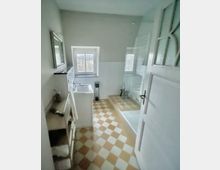 Helles Badezimmer mit weiß-beigem Schachbrettmuster-Boden, kleiner Fensteröffnung, Waschbecken mit Spiegel, Regalfach mit Handtüchern und einer ebenerdigen Dusche mit Glasabtrennung.