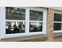 Außenansicht eines Klassenraums durch große, weiße Fenster mit Sprossen in einer Klinkerfassade. Im Raum sind Tische und Stühle symmetrisch angeordnet, die Fenster spiegeln Bäume und den Außenbereich.