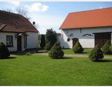 Ein ländlicher Hof mit einem Wohnhaus und einer Scheune, beide weiß gestrichen und mit roten Ziegeldächern. Dazwischen befindet sich eine gepflegte Rasenfläche mit kleinen Kegelformen geschnittenen Büschen und gepflasterten Wegen.