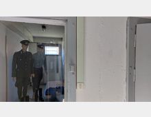 Ansicht eines Raumes mit einer Glaswand, hinter der zwei bekleidete Schaufensterpuppen in Uniform stehen. Der Raum hat weiße Wände, eine einfache Deckenleuchte und ein kleines vergittertes Fenster.