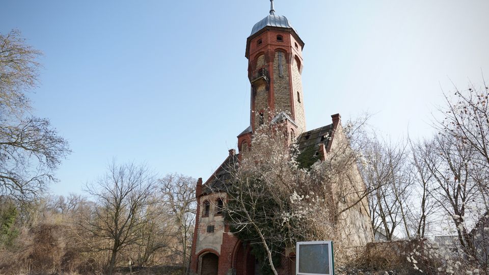 Historischer Backstein-Wasserturm mit achteckigem Schaft und Metallkuppel, an den ein kleines Gebäude mit Satteldach und Rundbogenportal anschließt. Der Turm steht in einer parkähnlichen Umgebung mit kahlen Bäumen und Sträuchern unter klarem Himmel.