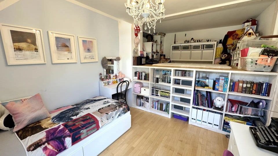 Helles Dachzimmer als Schlafzimmer mit Holzboden, weißem Bett und Schreibtisch sowie einem Kristall-Kronleuchter an der Decke. Entlang der Wand steht ein großes weißes Regalsystem mit Büchern, Boxen und Spielzeug, darüber ein erhöhtes Podest mit weiteren Aufbewahrungsmöbeln.