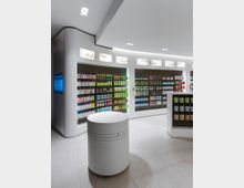 Innenraum einer modern gestalteten Apotheke oder Drogerie, mit beleuchteten Regalen voller Pflege- und Kosmetikprodukte. Im Vordergrund ein zylindrischer, weißer Tresen und eine helle, minimalistische Einrichtung.