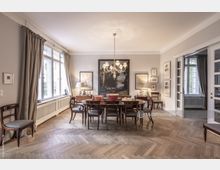 Esszimmer in einem klassischen Stil mit hölzernem Parkettboden, einem großen Esstisch aus Holz, umgeben von Stühlen mit gepolsterten Sitzflächen. Große Fenster mit beigen Vorhängen lassen Tageslicht herein, während ein Kronleuchter den Raum illuminiert.