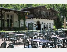 Außenbereich eines traditionellen Restaurants mit Biergarten, umgeben von Bäumen und bepflanzten Blumenbeeten. Im Hintergrund ist ein Gebäude mit Holzfassade und Blumenkästen an den Fenstern zu sehen, davor stehen zahlreiche leere Tische und Stühle.