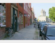 Straßenszene mit roten Backsteingebäuden auf der linken Seite, deren Fenster und Türen teils beschädigt sind, sowie geparkten Autos entlang der rechten Straßenseite. Im Hintergrund ist ein grauer Turm mit Zinnen und Spitzdächern zu sehen, umgeben von Bäumen.