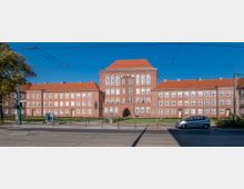 Backsteinbau mit symmetrischer Fassade und rot gedecktem Satteldach, bestehend aus einem zentralen Flügel mit Arkadeneingang und zwei Seitenflügeln mit zahlreichen weißen Sprossenfenstern; Straßenbahnkabel und eine Straße im Vordergrund.