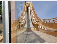 Holzstruktur mit einer großen, kurvigen Rutsche und seitlichen Geländern, die nach oben führt; eine Person mit Helm fährt auf einer der Bahnen hinunter. Im Vordergrund spiegelt sich die Anlage teilweise an einer Glasfläche.