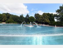 Ein großes Freibad mit klarem, blauem Wasser und mehreren Wasserspeiern. Im Hintergrund sind eine grüne Parklandschaft mit Bäumen und einige Besucher am Beckenrand erkennbar.