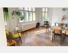 Heller Innenraum mit hohen Fenstern, einem Holzboden im Fischgrätmuster und einer schlichten Einrichtung. Der Raum enthält gelbe und grüne Sitzmöbel, einen weißen Heizkörper unter den Fenstern, Zimmerpflanzen und einen Esstisch aus Holz mit verschiedenen Stühlen.