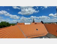 Blick über rote Ziegeldächer eines Gebäudekomplexes mit einem Kirchturm im Hintergrund, umgeben von Bäumen und einer weiten Landschaft unter einem Himmel mit weißen Wolken.