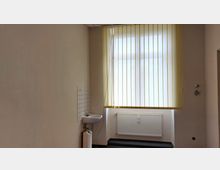 Ein schlichter Raum mit beigefarbenen Wänden, einem Fenster mit vertikalen gelben Jalousien, einem kleinen weißen Waschbecken an der linken Wand und einem weißen Heizkörper unter dem Fenster. Der Boden ist mit einem dunklen Teppichboden ausgelegt.