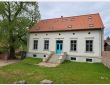 Zweistöckiges Wohnhaus mit heller Putzfassade und einem roten Ziegeldach, ausgestattet mit mehreren Dachfenstern. Vor dem Haus befindet sich eine Eingangstreppe mit türkisfarbener Tür, umgeben von einer Wiese; links steht ein großer, belaubter Baum.